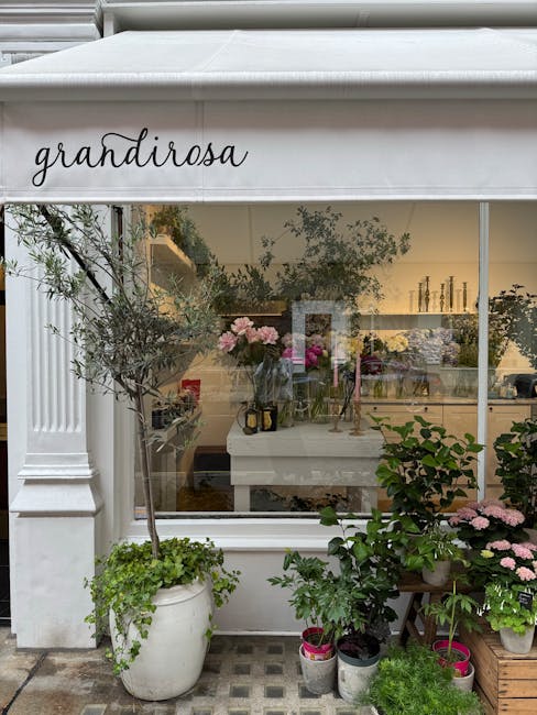 The image shows the exterior of a flower shop named 'grandiorosa' with large glass windows revealing an interior filled with fresh floral arrangements. In front of the shop, there are several potted p